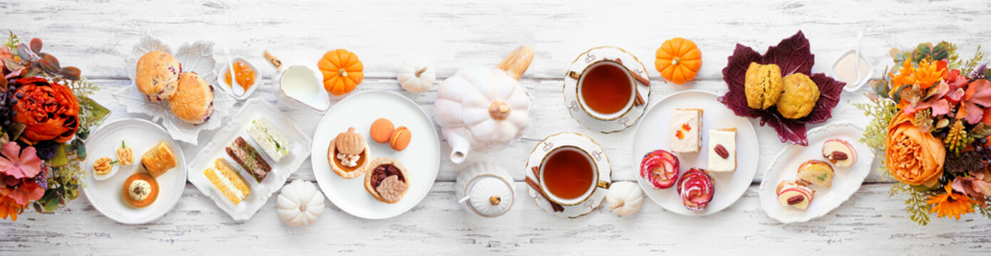 Autumn theme afternoon tea table scene over a white wood banner background. Pumpkin and apple theme savory finger foods, sandwiches, scones, and desserts. Above view.