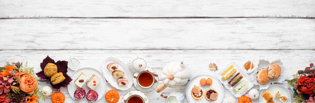 Autumn theme afternoon tea bottom border against a white wood banner background. Pumpkin and apple theme savory finger foods, sandwiches, scones, and desserts. Top down view with copy space.