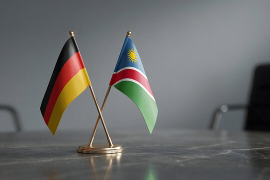 Miniature desk flags of germany and namibia crossed on a metallic stand over a dark marble surface with a soft grey background, in an office