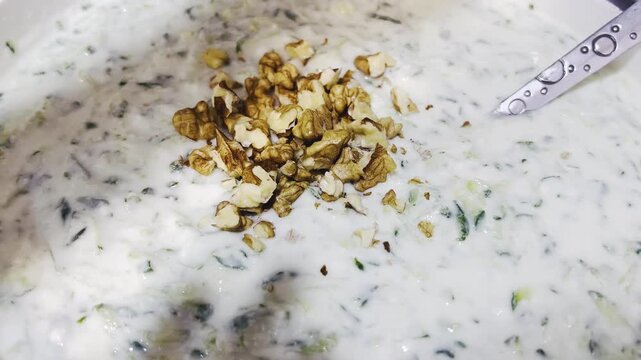 Adding crushed walnuts to Persian yogurt cucumber dip close up 16:9, homemade appetizer concept