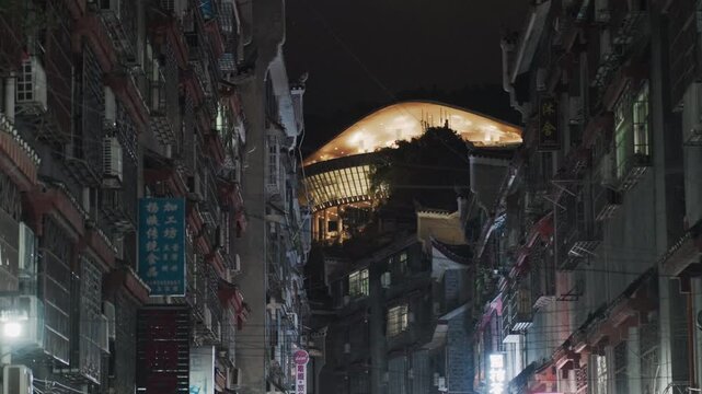 Night alley under glowing moon, narrow wet street framed by tall apartment facades, neon signs and hanging laundry, dim lamplight casting long shadows, puddles reflecting illuminated dome and shop