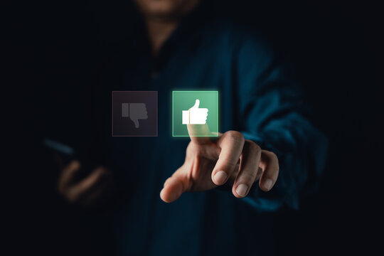 Man pointing finger to click glowing virtual thumbs up icon on dark background. Concept of customer satisfaction, positive feedback, online review, excellent service, and approval.