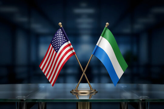 Small crossed flags of the united states of america and sierra leone displayed on a glass table in a dark blurred office interior setting