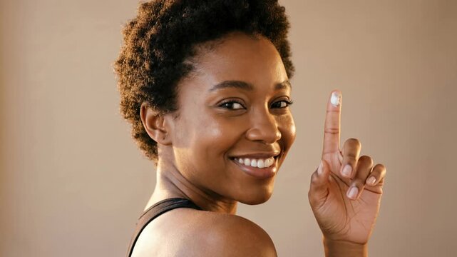 Happy Black woman smiling with a dab of skincare cream on her index finger while winking against a warm neutral studio backdrop