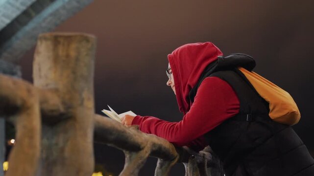 Night woman headscarf leaning on railing, orange backpack and phone, city light glow, wooden posts in foreground, candid side profile, layered winter clothing, quiet contemplative mood, cinematic
