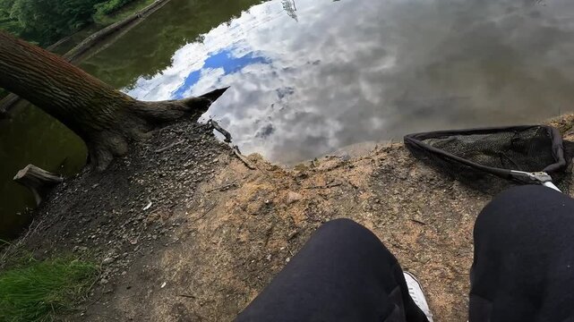 Point of view of seated angler looking at fishing rods set up on bank by calm forest lake, relaxing summer hobby concept.