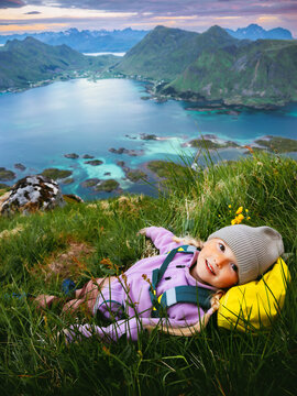 Tourist child travel in Norway girl laying on the grass enjoying Lofoten islands sea and mountains view, family summer vacations outdoors happy kid backpacker healthy lifestyle candid moments