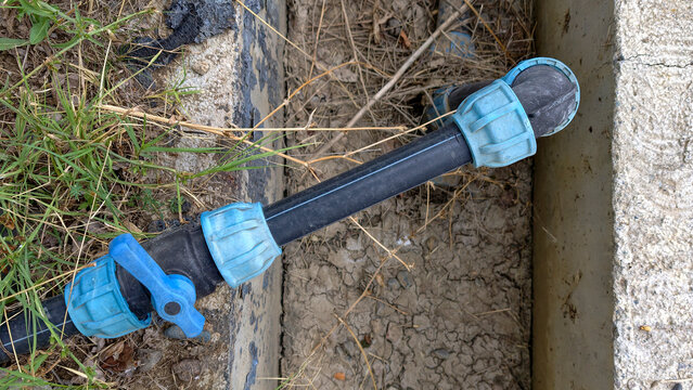 Black polyethylene pipe with blue compression fittings and a valve connecting sections. Laying on dry. Cracked soil next to a concrete edge