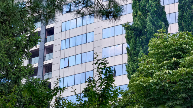 Modern architectural building with a reflective glass facade and multiple balconies. Framed by vibrant green trees