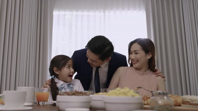 Happy Asian family relationships at the dining table in the morning, with father in a suit, mother, and a child girl smiling, teasing, and bonding before eating breakfast together in kitchen at home. 