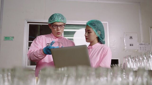 Two Asian quality control inspectors in uniform standing, using a laptop computer to analyze and inspect empty glass beverage bottles on moving conveyor belt in production line at industrial factory. 