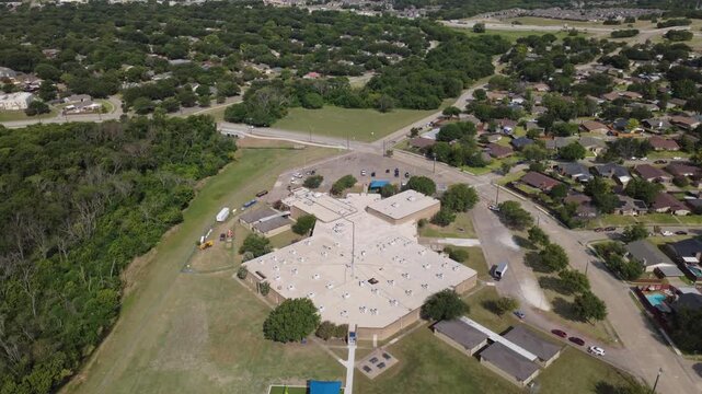 Detailed aerial view showcases flat roof structure of public middle school in Lyons Crest Estates in suburbs DFW. Tree lined forest buffers border school grounds, Garland, North Texas
