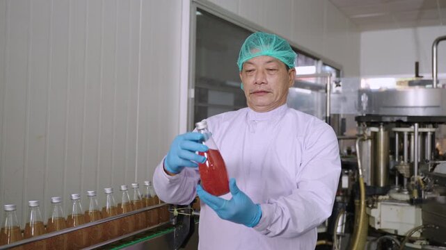Professional Asian inspector in uniform of food and drink factory holding a juice beverage bottle promoting and advertising healthy nutritional benefits in production line at a manufacturing industry.