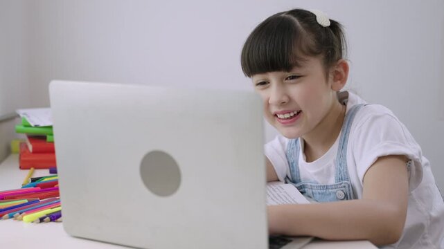 Cute Asian girl student sitting at desk using a laptop computer for studying online learning via video calling with remote teacher in a home school. Homeschooling education and new normal concept.
