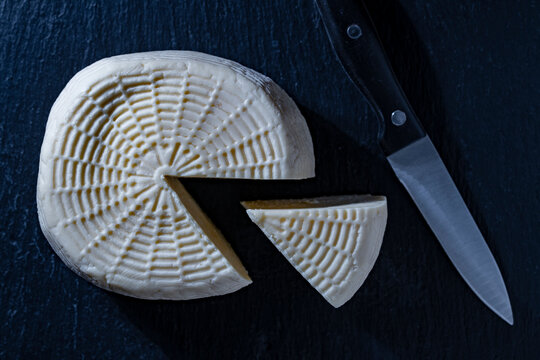 Fresh traditional homemade cheese with knife on dark background, closeup. Top view of round head of artisanal cheese with cut wedge and knife on black textured background