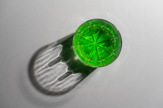 Glass of bright green soda drink with beautiful shadow on white surface, closeup. Top view of crystal glass filled with carbonated green liquid casting dramatic caustic shadow on light background