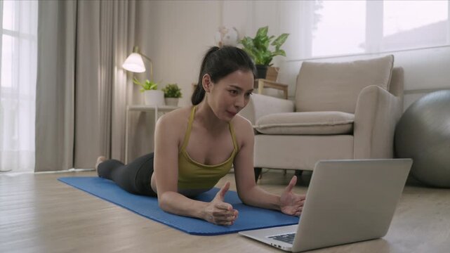 Happy Asian young woman yoga instructor teaching, talking, and video chatting on laptop webcam camera in plank position on a mat for healthcare in living room at home. Fitness training class concept. 