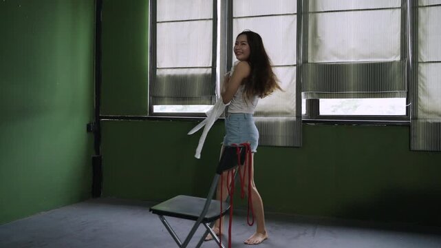 Unhappy and psychopathic Asian woman with unusual mental health issues is dancing alone pretending to be happy with red rope and chair in her room. Lonely female is stressed and about a failed life.