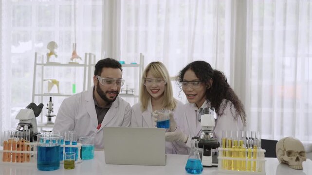 Group of happy men and women ethnic diversity, scientists and researchers wearing lab coats and goggles sitting and using a computer laptop for an online conference meeting together in the laboratory.