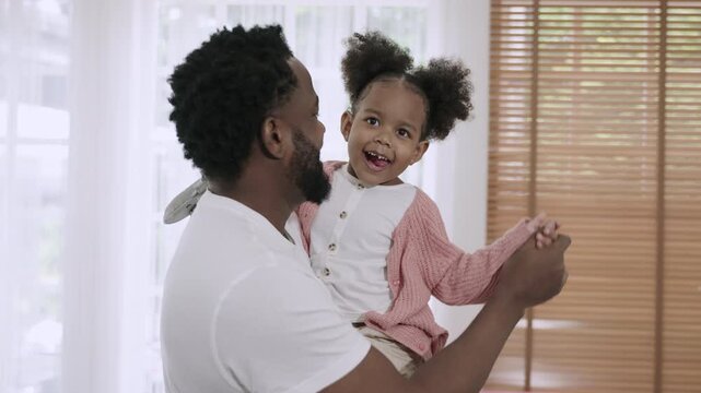 Happy African American father standing smiling and holding his cute little daughter while dancing together with love and bonding, having fun and cheerful in living room at home. Father's Day concept. 