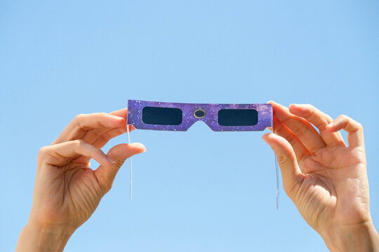 Woman hands holding purple solar eclipse glasses against blue sky background. Solar eclipse glasses are specially certified, ultra-dark glasses to protect eyes from damage when looking directly at sun