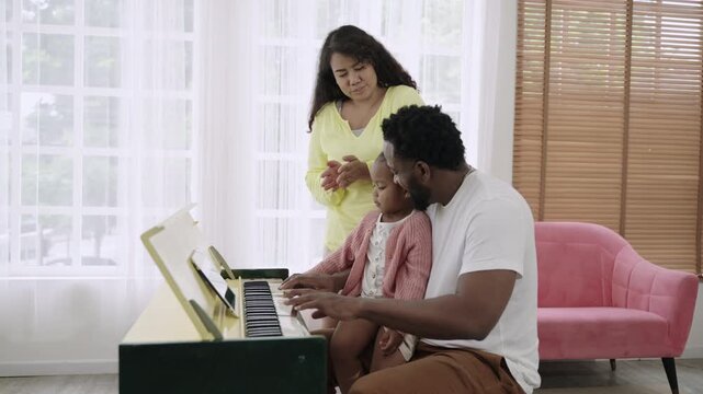 Happy African American family playing piano together with fun and cheerfulness in the living room at home. Black father teaching his cute little daughter to play the piano with love and bonding. 