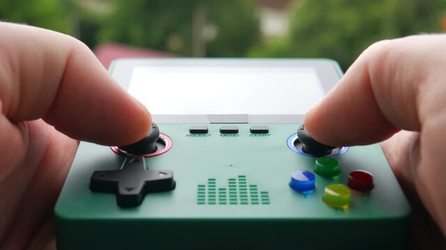 Close-up of female gamer hands holding and playing a green retro portable video game console outdoors against a blurred nature background.