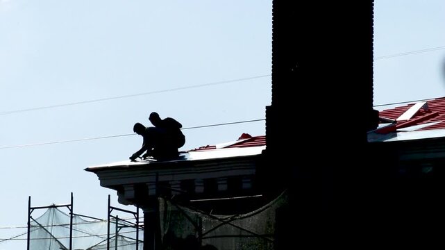 Two construction workers repairing red metal roof on building silhouette against blue sky