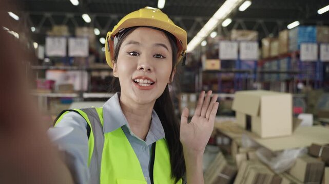 Happy Asian young woman warehouse worker in uniform wearing a yellow safety helmet, standing smiling and holding a camera while talking and greeting via video call in a logistics distribution factory.