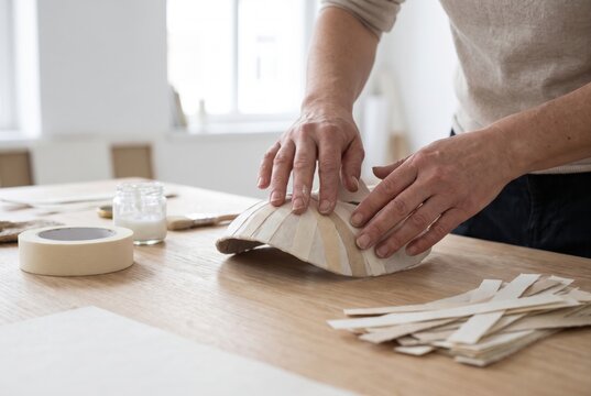 Hands applying strips of paper to a curved mold while making a papier mache object on a wooden crafting table