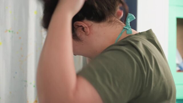 Novice looms over clay workspace. Apprentice in denim jeans and green shirt settings up materials. Trainee carefully arranges teal straps and apron in busy ceramic studio environment