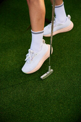 Obraz premium Close up of woman feet wearing stylish white athletic shoes on mini golf turf. Female player holding silver putter on green synthetic grass
