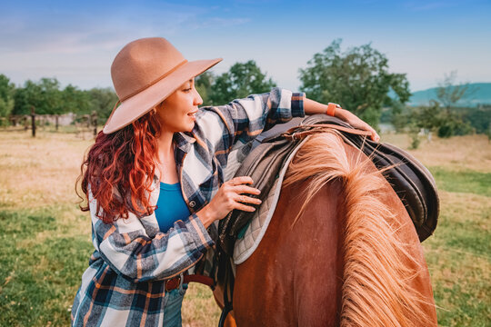 Young woman adjusting leather saddle on chestnut horse outdoors before a ride.