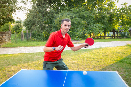 Young man in a red shirt raises a ping pong paddle mid-game at an outdoor tennis table, ball blurred in motion. Active leisure concept.