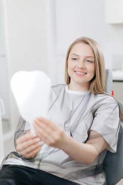 Young caucasian female patient smiling at dental clinic holding mirror, white light