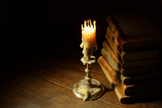 A candle stands on a silver holder, casting light on a stack of old books next to it.  