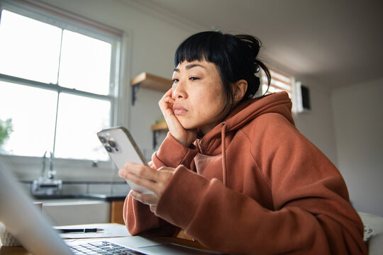 Woman using smartphone and laptop at home kitchen table
