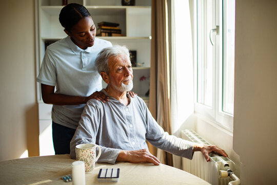 Caregiver comforting elderly man at home by window radiator