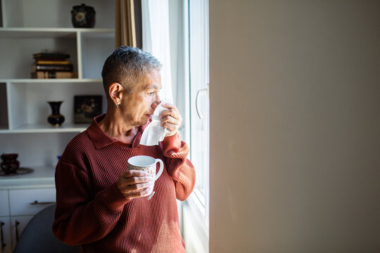Older adult with tissue and tea looking out window at home
