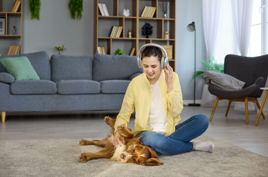 Woman and dog cuddling at home with headphones. Smiling owner enjoys music while her spaniel lies on the rug, soaking up affection in a living room. Warm, bonding human pet connection.