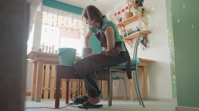 Female potter shaping clay pottery workshop, sunlit studio space, shelves of pots and plants, bucket on table, stool and chair, artist wearing apron leans forward, hands covered in wet clay, careful