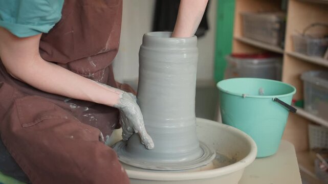Pottery workshop caucasian apprentice shaping clay on wheel, forming tall cylinder beside turquoise bucket, wet hands in brown apron, focused slow craft atmosphere, tactile texture and steady motion