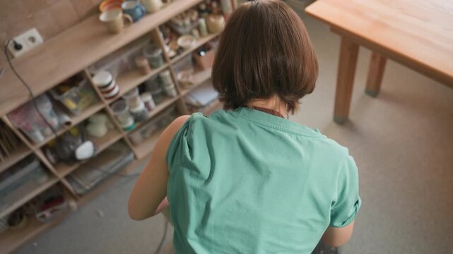 White woman student practicing wheel technique from overhead, green shirt and apron visible, bucket and shelves of bisque ware, classroom mood focused on learning and steady handwork