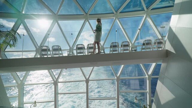 A woman in a hat walks across a glass-floored observation bridge inside a modern cruise liner in the Caribbean Sea. Sunlight streams through geometric windows with ocean wake visible below.