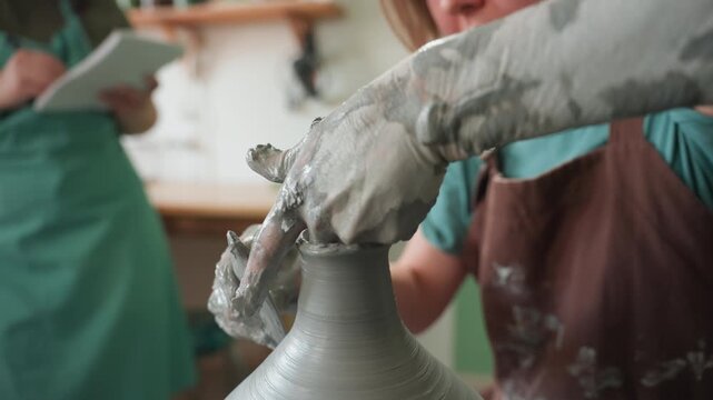 Caucasian potter fingers shaping clay rim on wheel in pottery workshop, closeup tactile shot of wet hands and fingers, observer with notebook in background taking notes, focused technique, smoothing