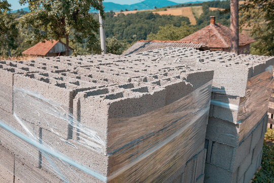 Pallets of hollow concrete blocks wrapped in plastic sheeting stacked outdoors in warm summer light, ready for a rural construction project.