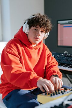 Young male music producer using a hardware controller in a studio wearing a red hoodie and headphones for beatmaking background and social media content