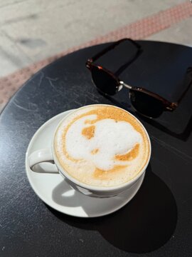 Top view of a white cappuccino cup with imperfect latte art on black outdoor table, with black sunglasses. No people, clean space, lifestyle ugc photo concept for social media