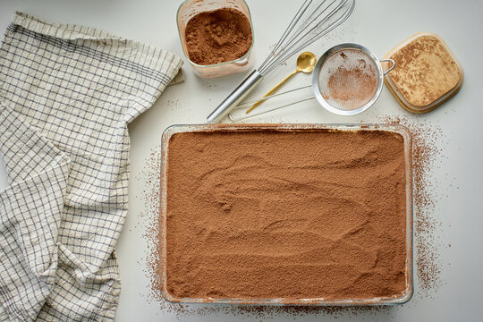 Cocoa topped tiramisu in glass dish on table, with sieve, whisk, cocoa bowl, spoon and towel