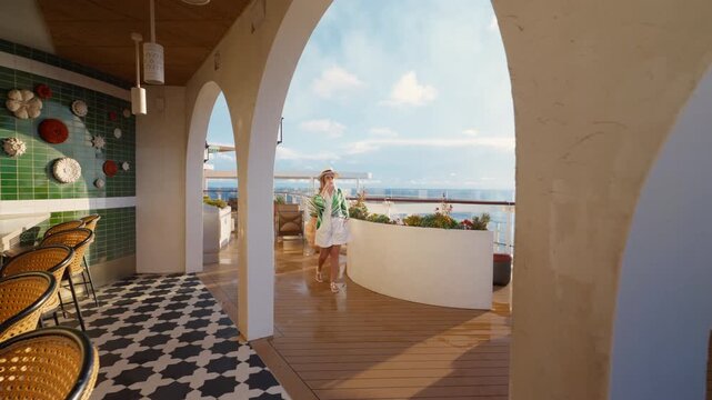 Elegant woman wearing straw hat and palm print kimono walks through luxurious cruise ship lounge featuring white Mediterranean arches, green tiled bar wall with decorative plates.